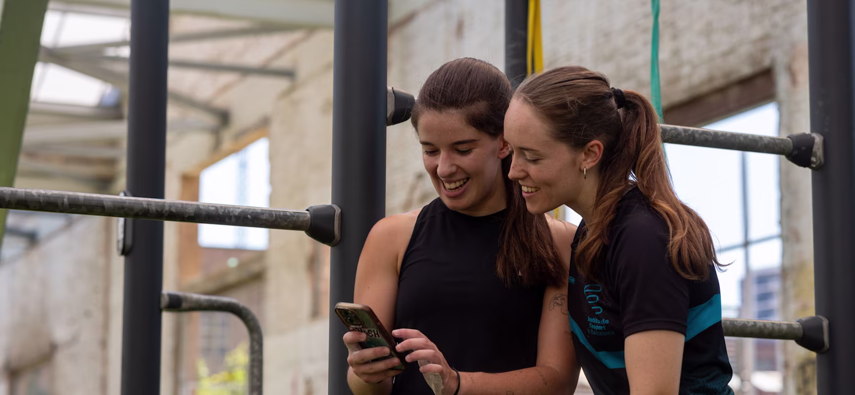 Two women at an outdoor gym stand beside metal bars, one holding a smartphone while the other leans in to look at the screen.
