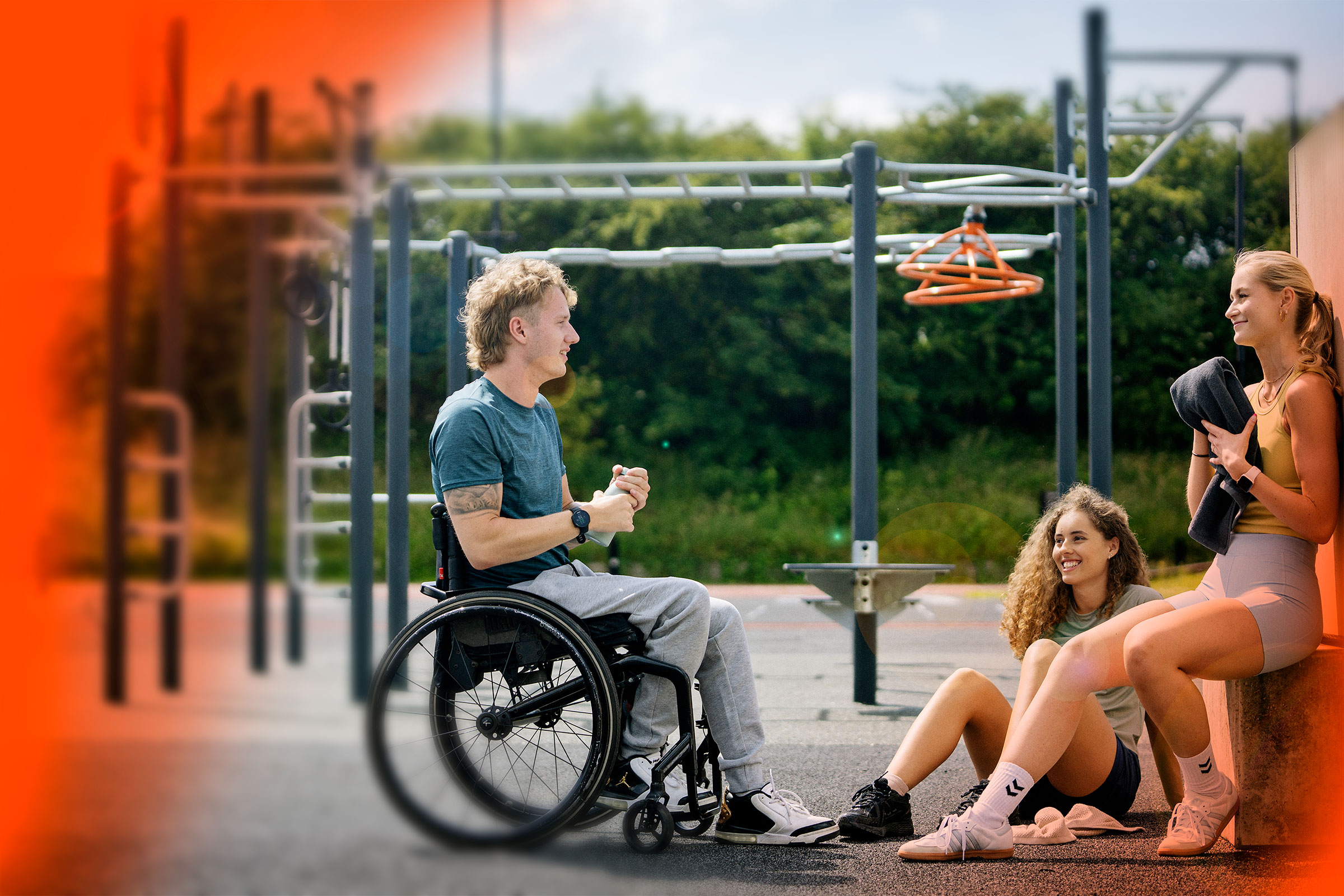 Man in a wheelchair chatting with two women resting near outdoor gym bars on a sunny day