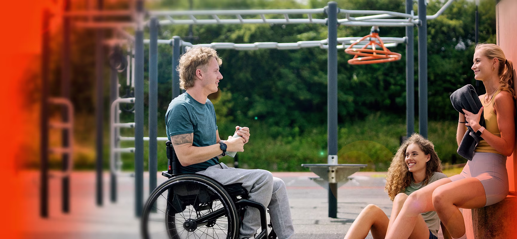 Man in a wheelchair chatting with two women resting near outdoor gym bars on a sunny day