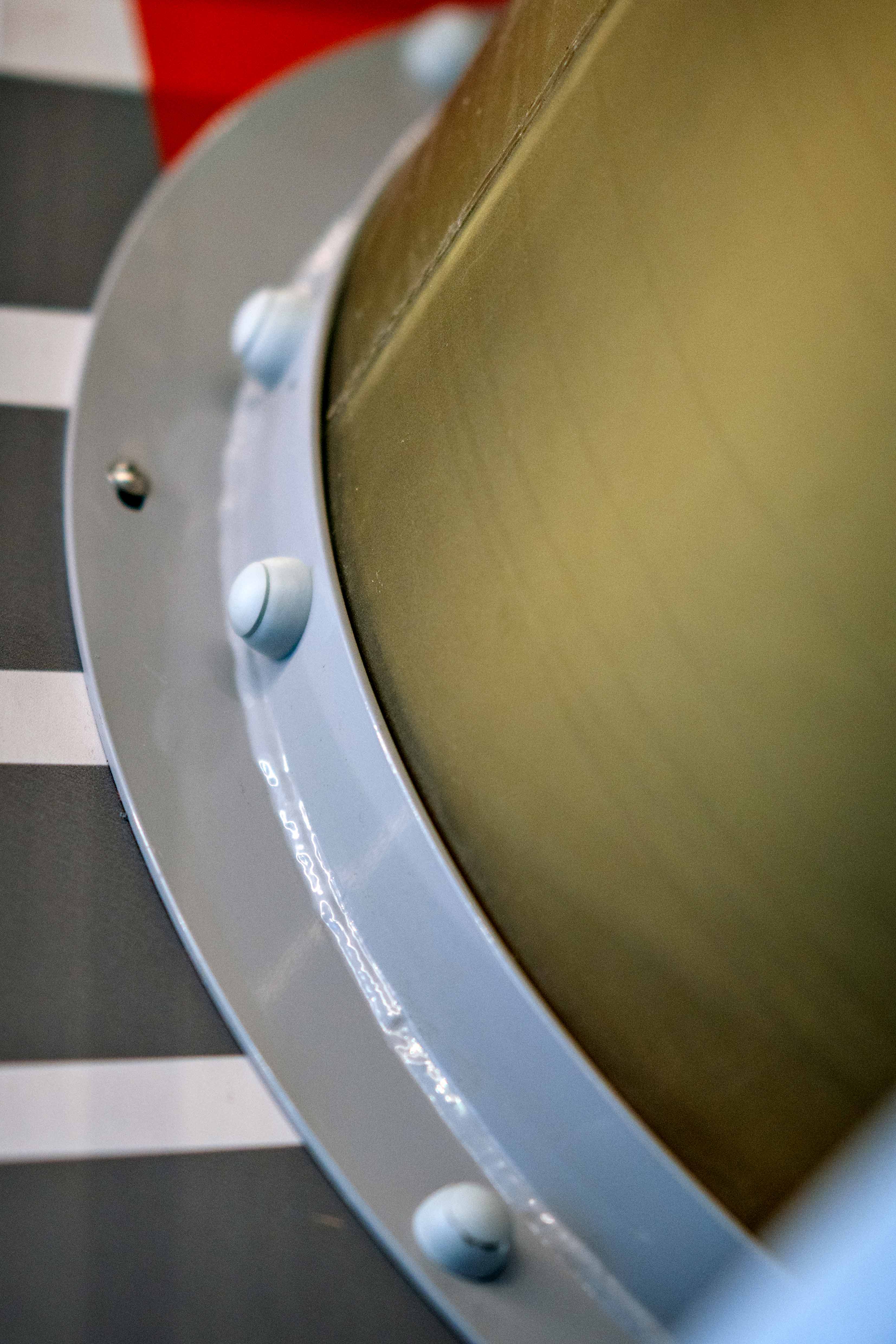 Close-up of a cylindrical metal structure with bolts on a pale blue ring, against striped grey and red background.