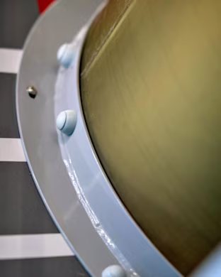 Close-up of a cylindrical metal structure with bolts on a pale blue ring, against striped grey and red background.