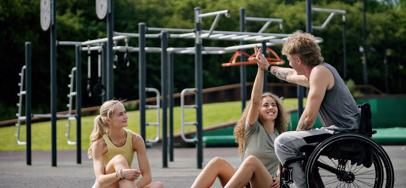 Three young athletes relax on an outdoor track; one in a wheelchair high-fives a seated teammate while another sits nearby smiling