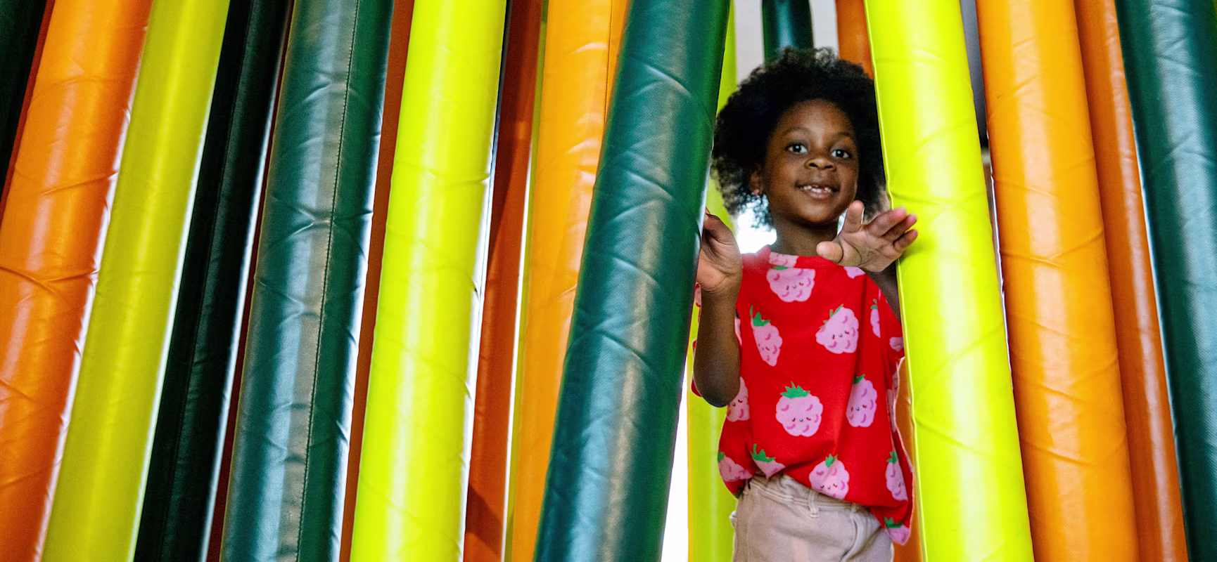 Child in red strawberry shirt walking through hanging padded tubes in bright yellow, green, and orange at an indoor play area