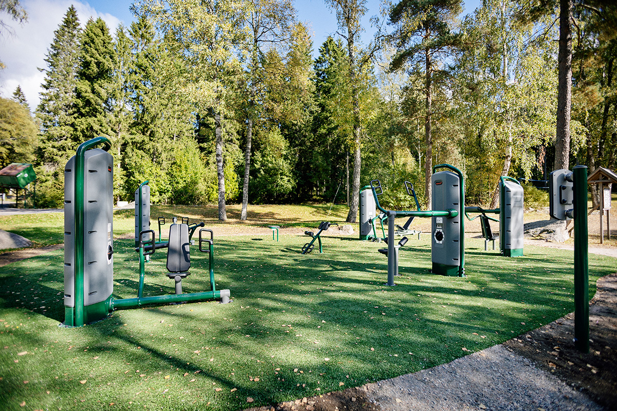 Outdoor gym with green and grey exercise machines on artificial grass, surrounded by tall trees in a sunny forest park.