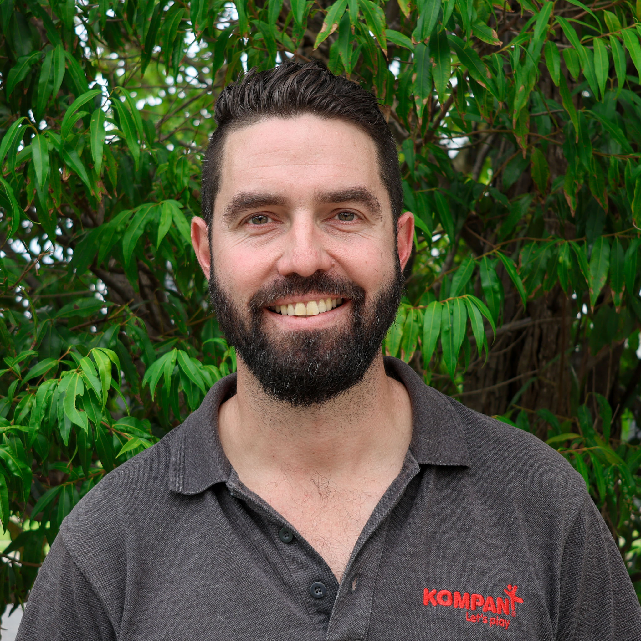 Person with blurred face wearing grey Kompan polo shirt, standing outdoors in front of leafy green trees.
