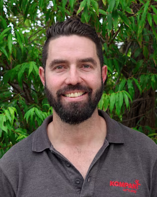 Person with blurred face wearing grey Kompan polo shirt, standing outdoors in front of leafy green trees.
