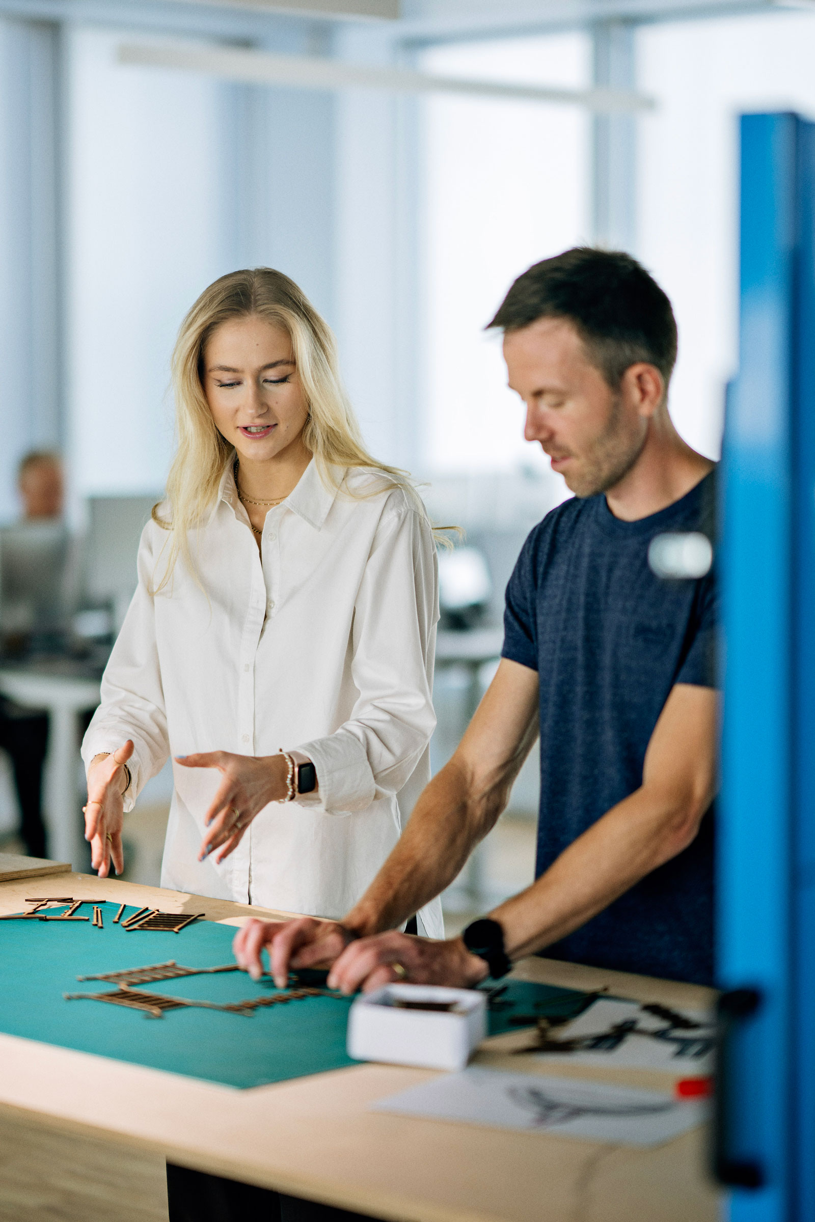 Two colleagues in a bright office lean over a workbench, arranging small wooden pieces on a green mat while discussing a design.