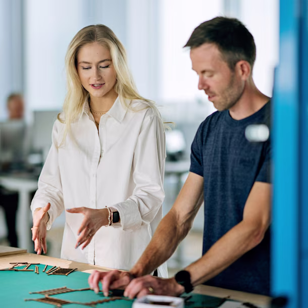 Two colleagues in a bright office lean over a workbench, arranging small wooden pieces on a green mat while discussing a design.