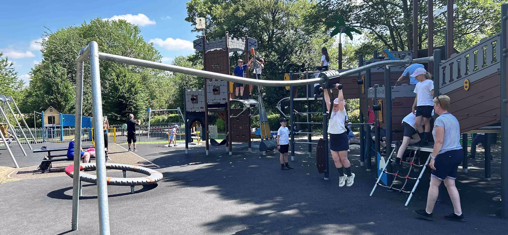 Children play on a pirate ship playground and swings in a sunny park, with adults nearby supervising under a blue sky.
