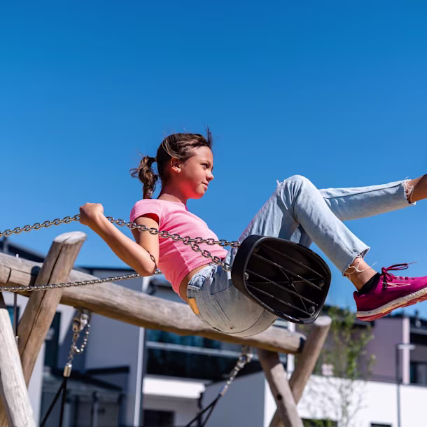 Blurred-face child in pink shirt and jeans swinging high on a wooden swing set against a clear blue sky near modern houses