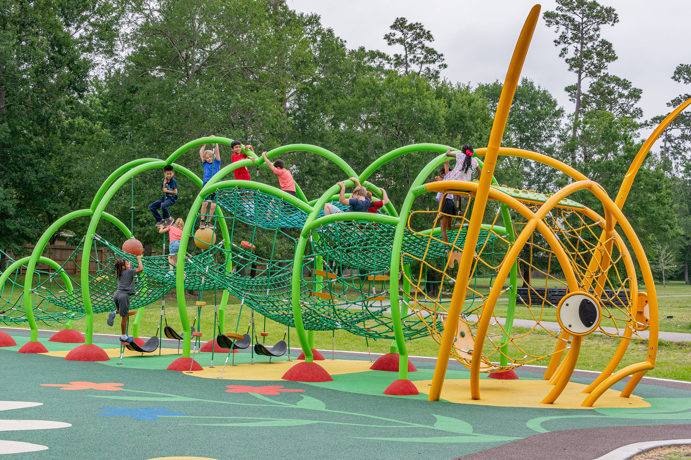 Children climb and hang on bright green and yellow rope playground structures in a park surrounded by tall trees.