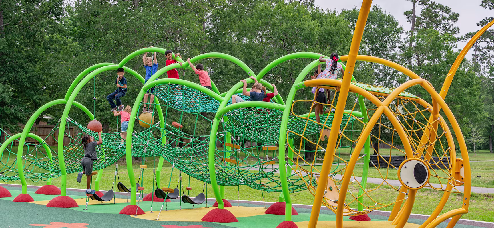 Children climb and hang on bright green and yellow rope playground structures in a park surrounded by tall trees.
