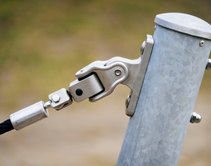 Close-up of a metal cable tensioner attached to a galvanized post outdoors, showing bolts and hinge mechanism.