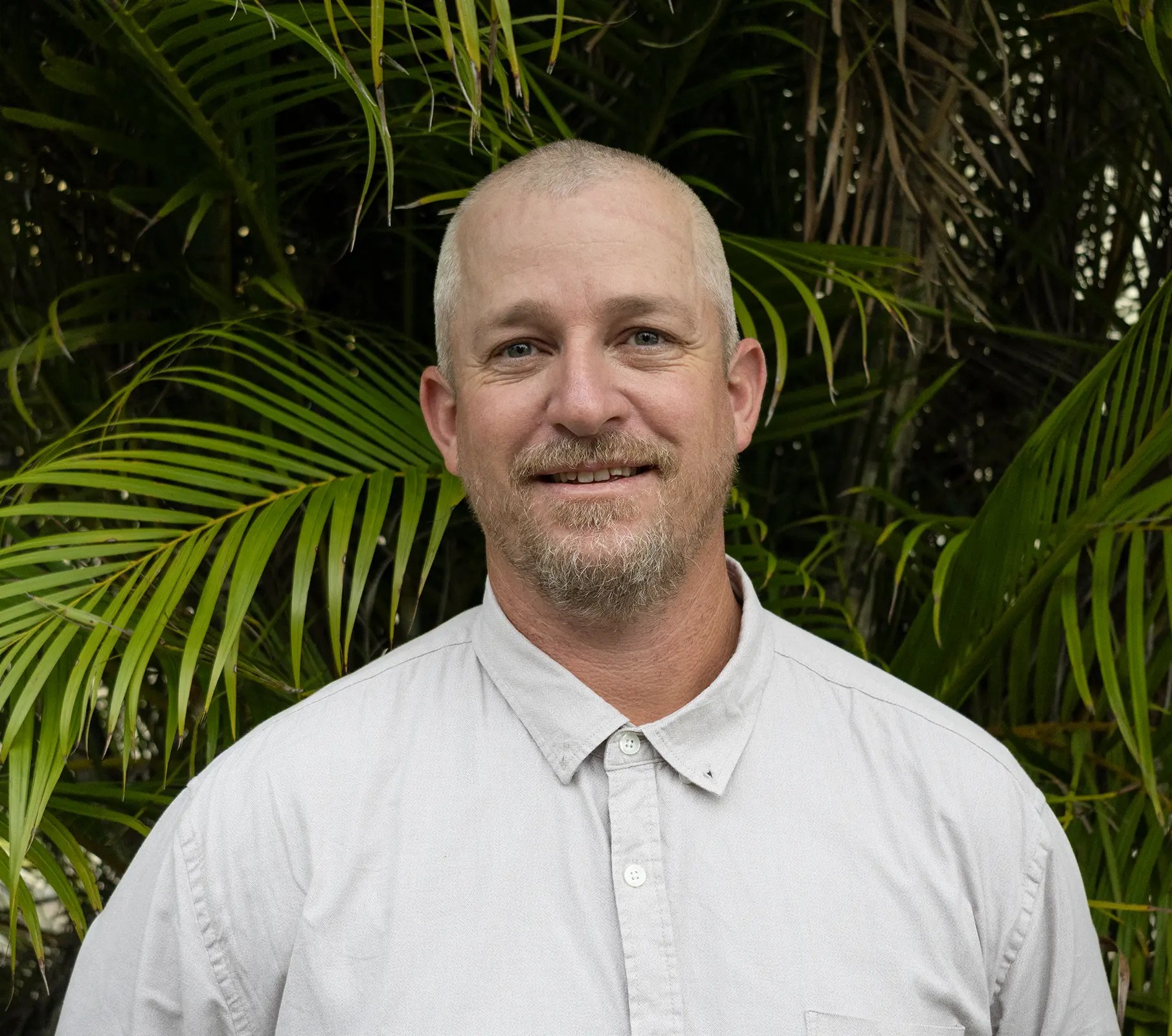 Portrait of a person with a blurred face, short hair, and dark clothing, standing outdoors in front of green leafy trees.