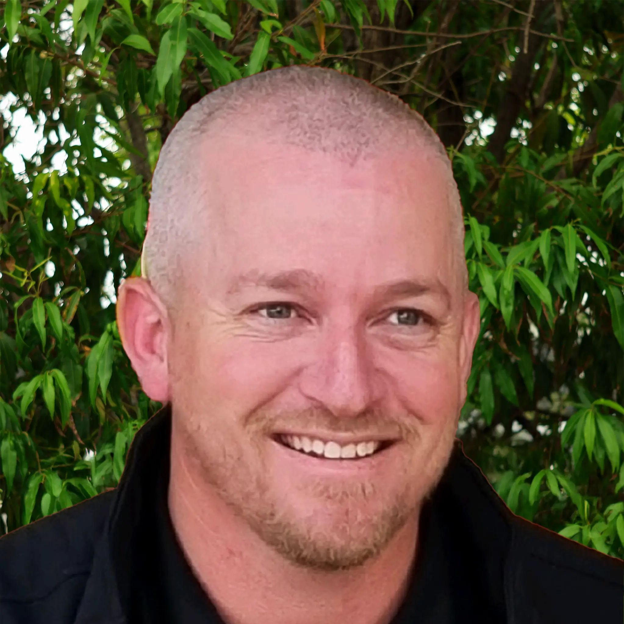 Portrait of a person with a blurred face, short hair, and dark clothing, standing outdoors in front of green leafy trees.