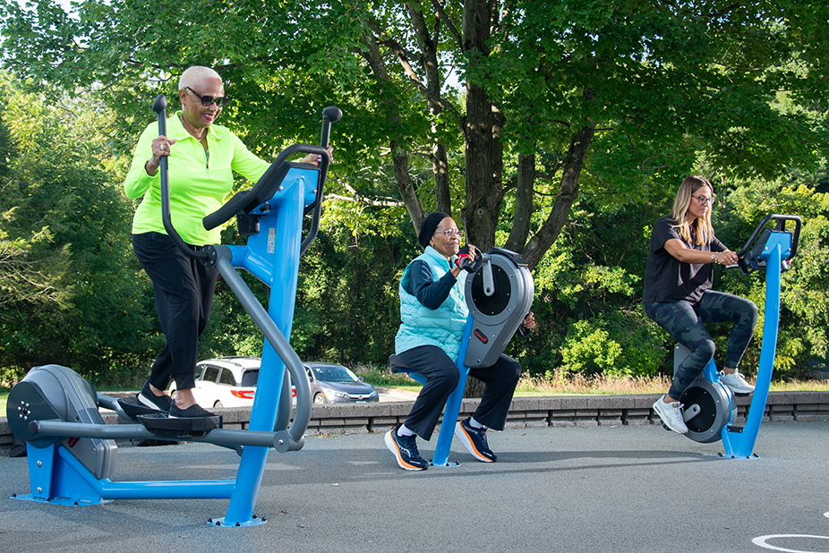 Three women exercising outdoors on commercial cross trainer, commercial exercise bike, and seated arm bike from KOMPAN.