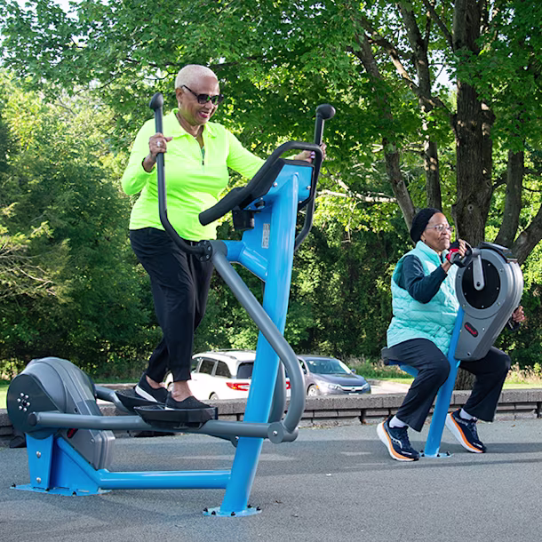 Three women exercising outdoors on commercial cross trainer, commercial exercise bike, and seated arm bike from KOMPAN.