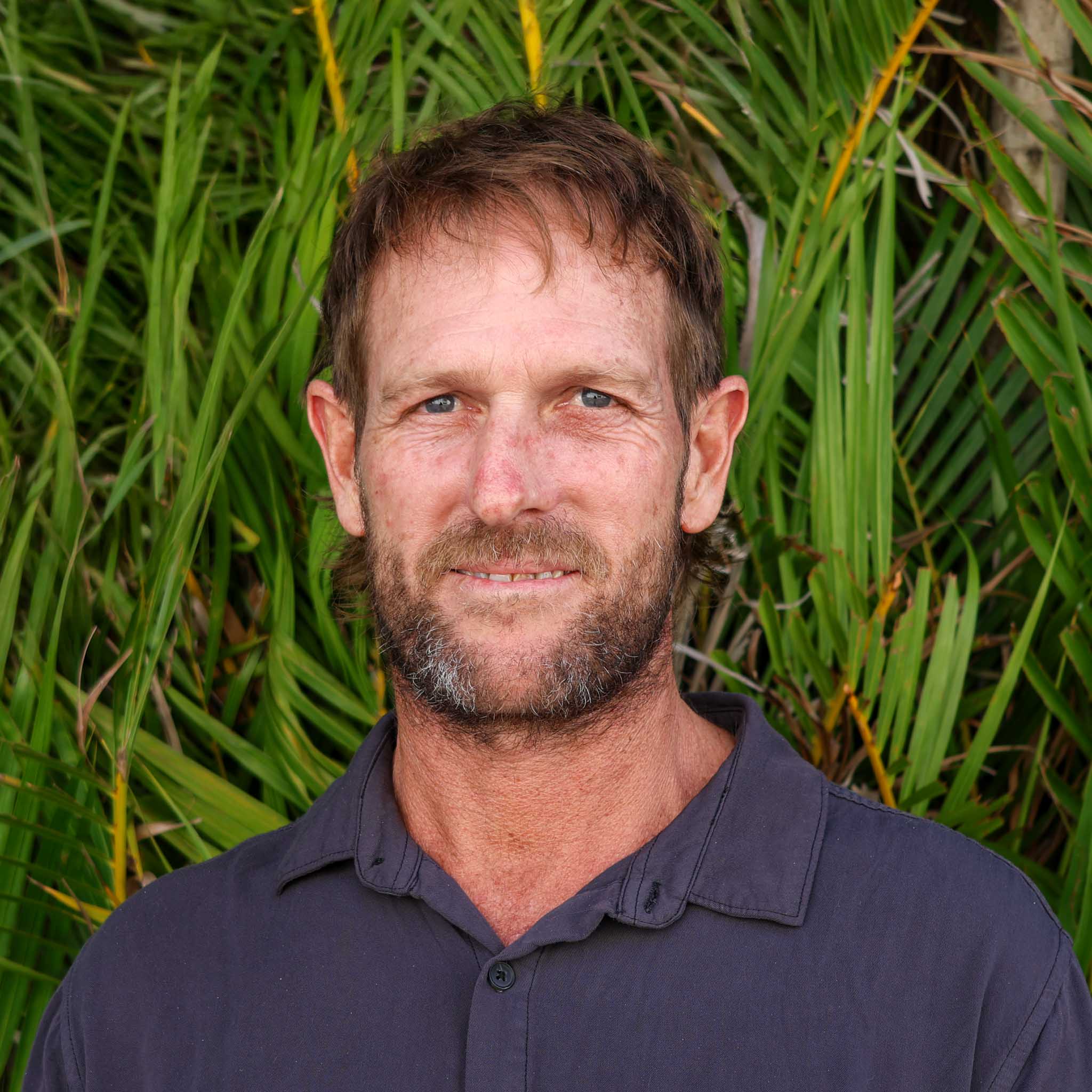 Person in a dark shirt standing in front of dense green tropical plants, with their face obscured by a blurred rectangle.