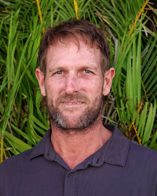 Person in a dark shirt standing in front of dense green tropical plants, with their face obscured by a blurred rectangle.