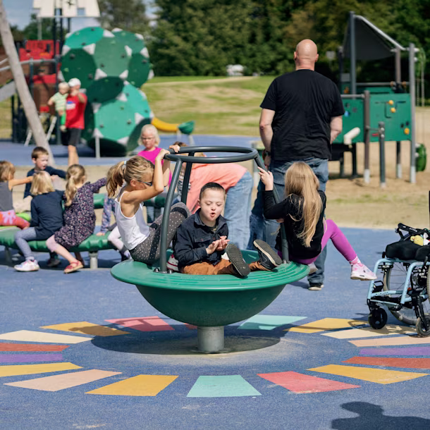 Children play on an inclusive roundabout in a colourful playground, with a wheelchair parked nearby and adults watching in the background