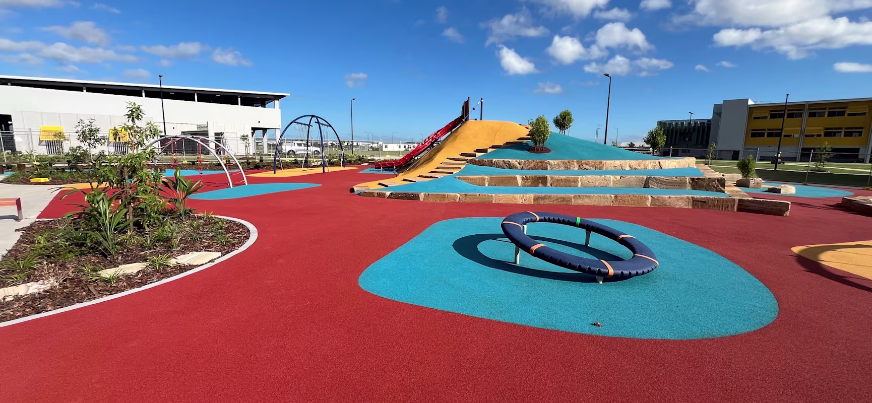 Colorful modern playground with red and blue rubber paths, climbing frames, and a slide on a grassy hill under a bright blue sky