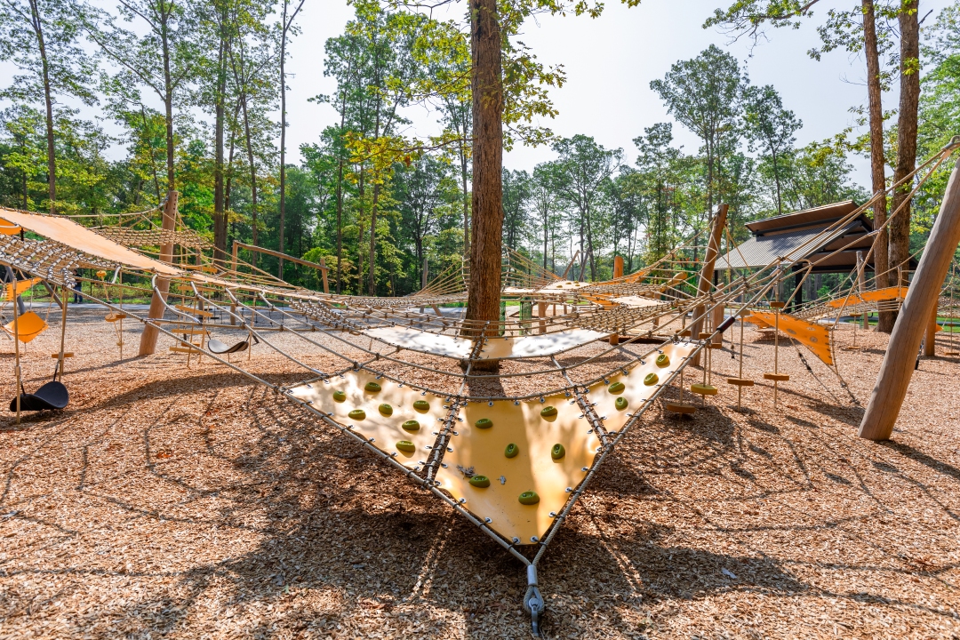 Rope and net climbing playground with yellow panels and wood-chip ground, set among tall green trees on a sunny day.
