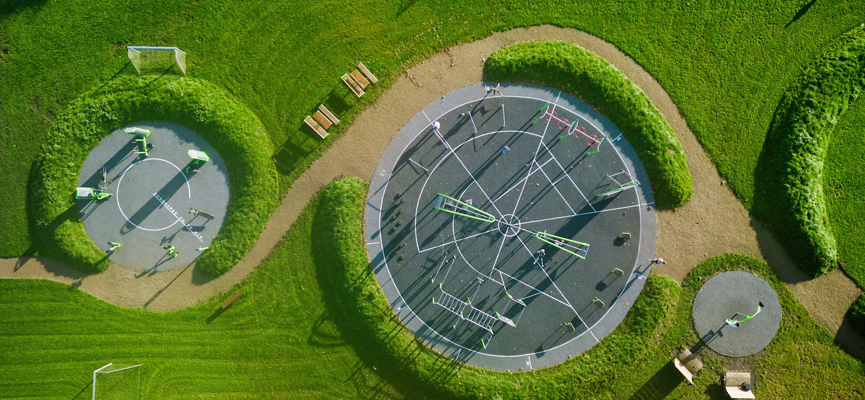 Aerial view of a green park with circular playgrounds, sports courts, paths, benches and scattered people using the equipment