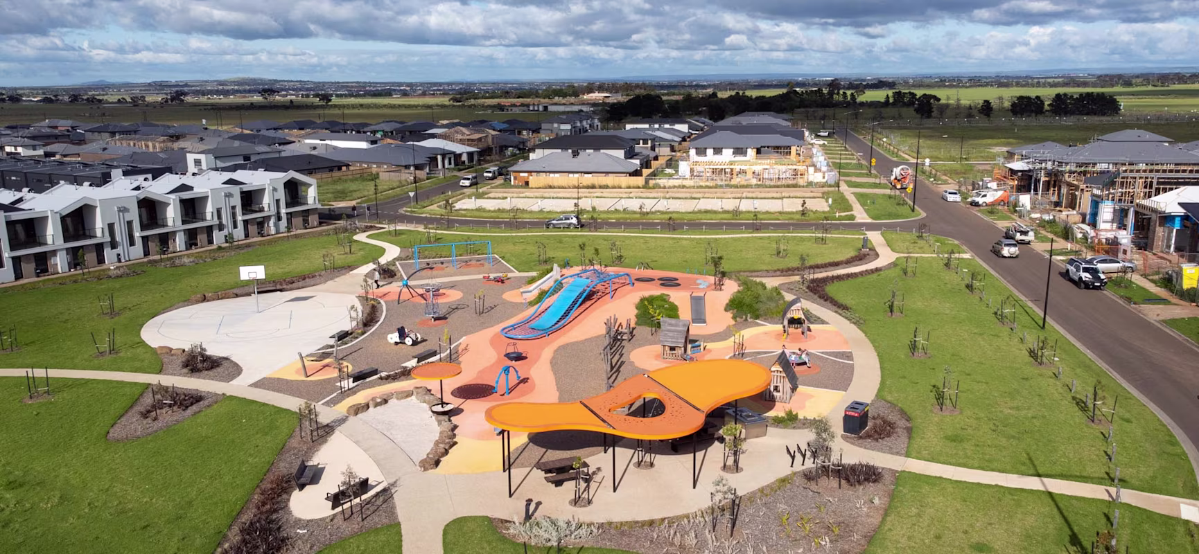 Aerial view of a colourful playground with orange shade sails in a green park, surrounded by new houses and a road under cloudy sky