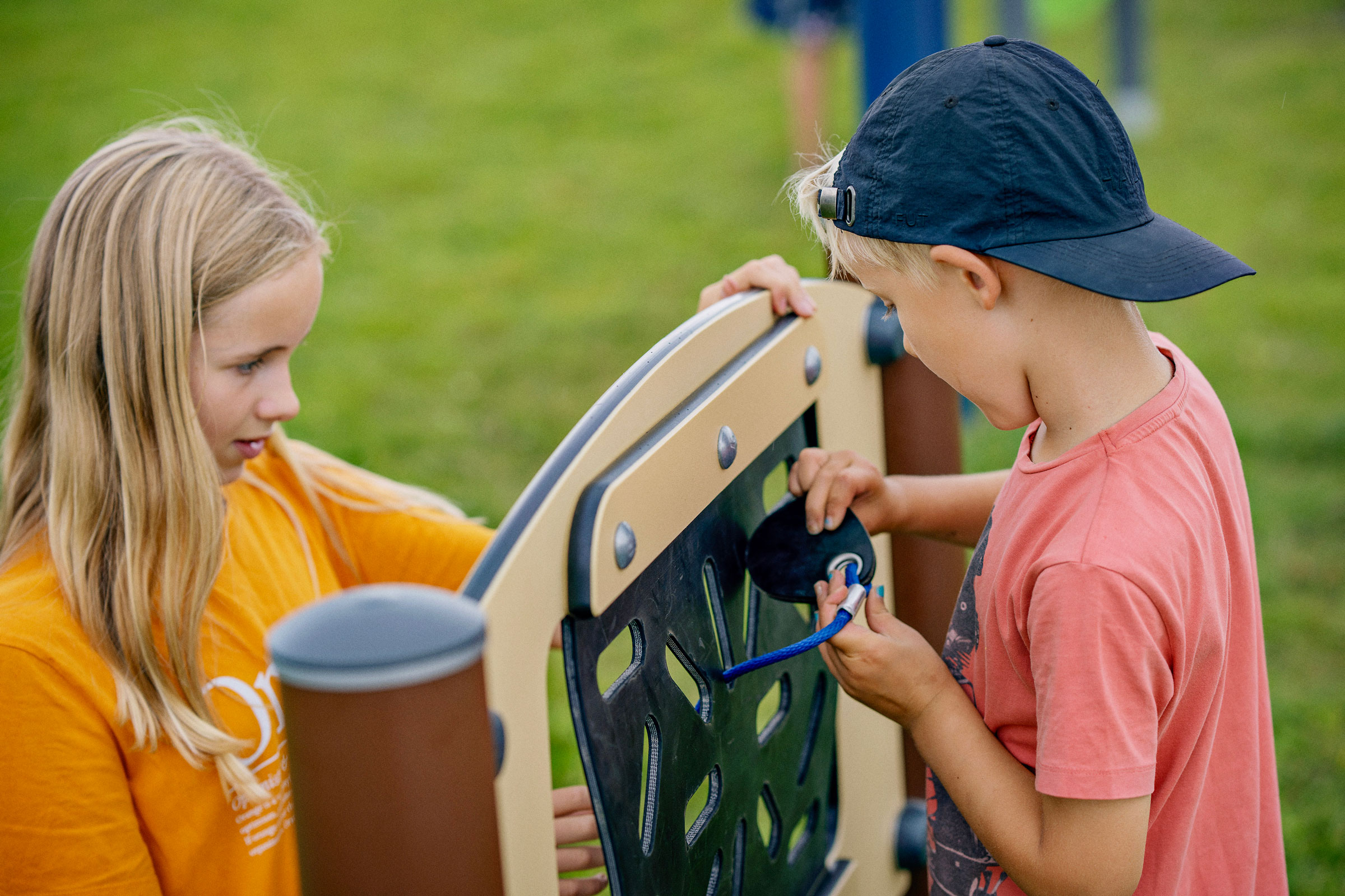 Two children at a playground panel, one in orange shirt, one in pink shirt and cap, threading a blue rope through holes.