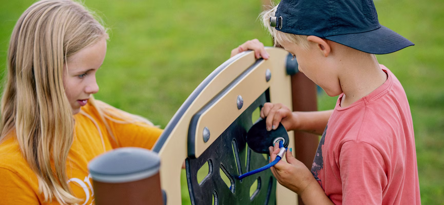 Two children at a playground panel, one in orange shirt, one in pink shirt and cap, threading a blue rope through holes.