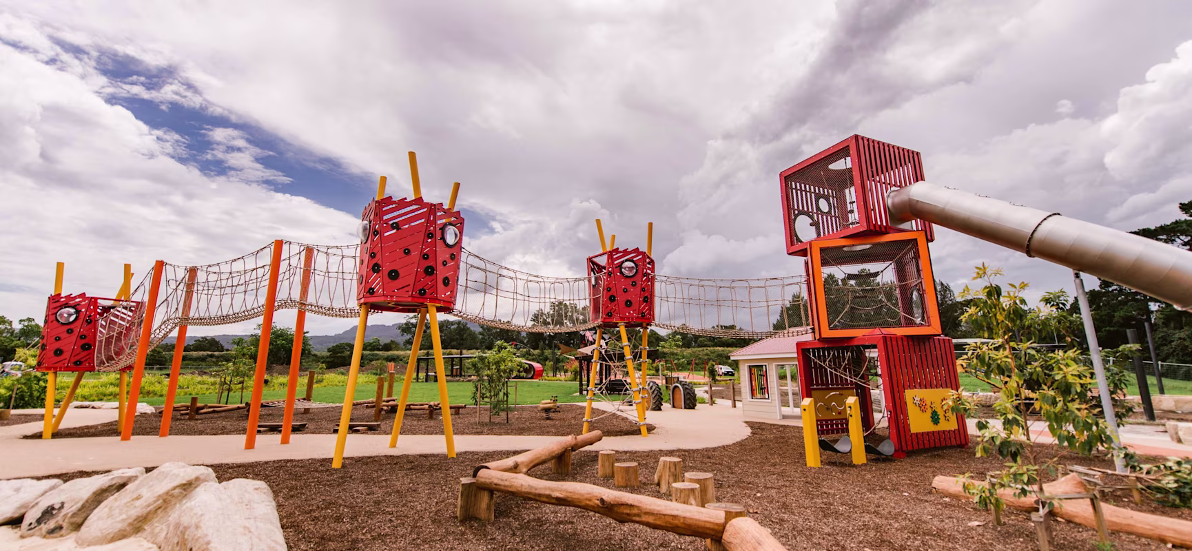 Outdoor playground with bright red climbing towers, rope bridges, and a metal slide, set in a park under a cloudy sky