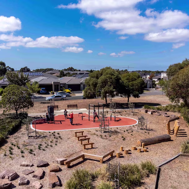 Small suburban park with red play area, people exercising on equipment, wooden logs and benches, trees and houses under blue sky