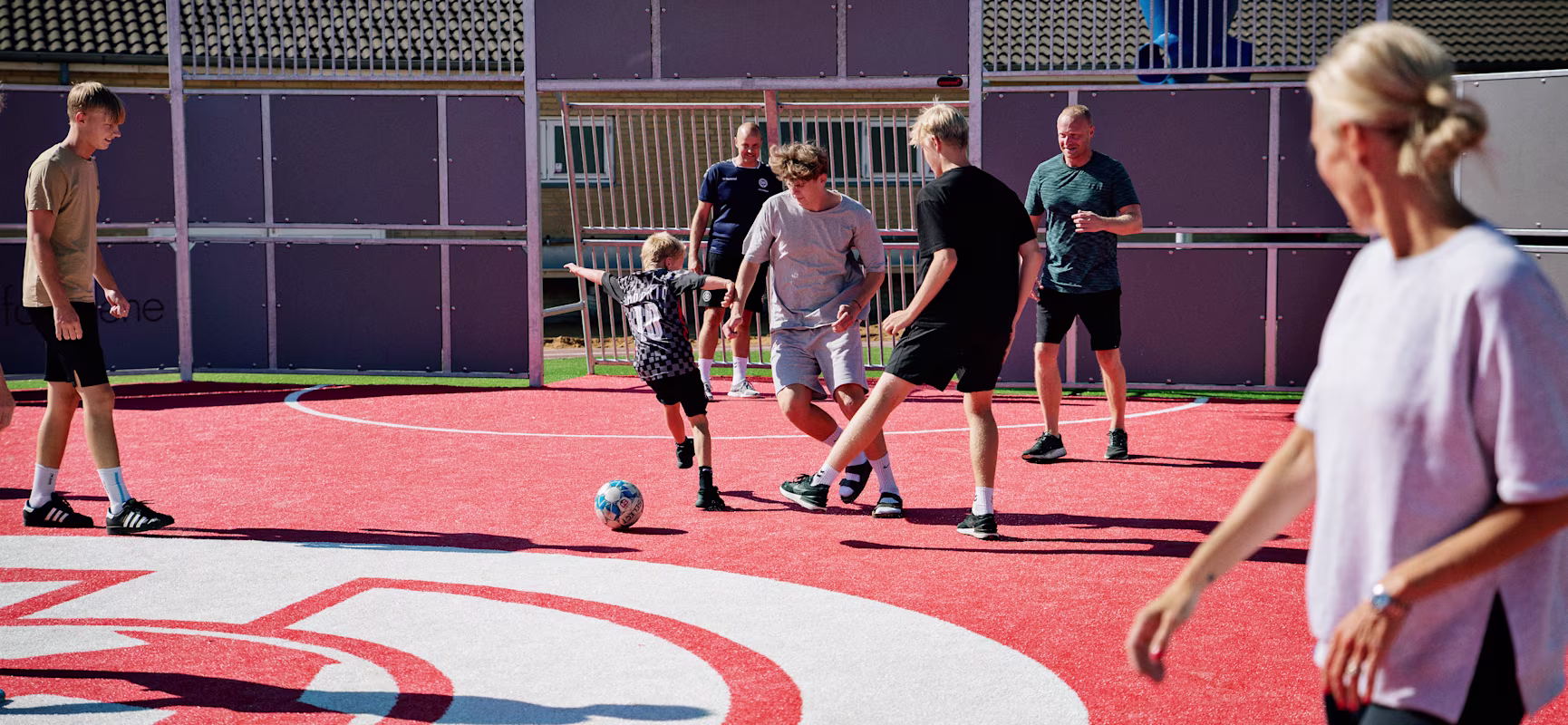 Group of children and adults playing football on a bright red outdoor court under a basketball hoop on a sunny day