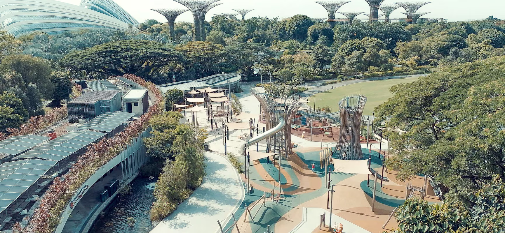 Aerial view of a modern playground among lush trees, with curving paths and futuristic tree-like towers in a large urban garden.