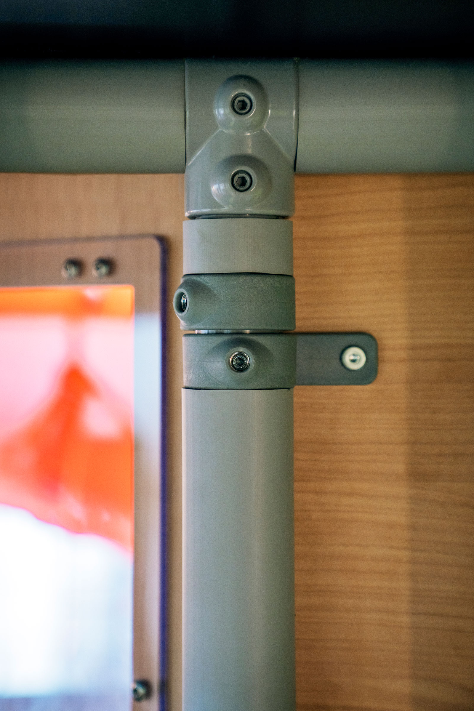 Close-up of grey metal pipes joined with brackets and screws against a wooden wall, with a blurred orange window on the left.