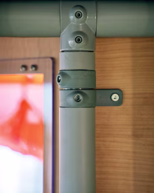 Close-up of grey metal pipes joined with brackets and screws against a wooden wall, with a blurred orange window on the left.