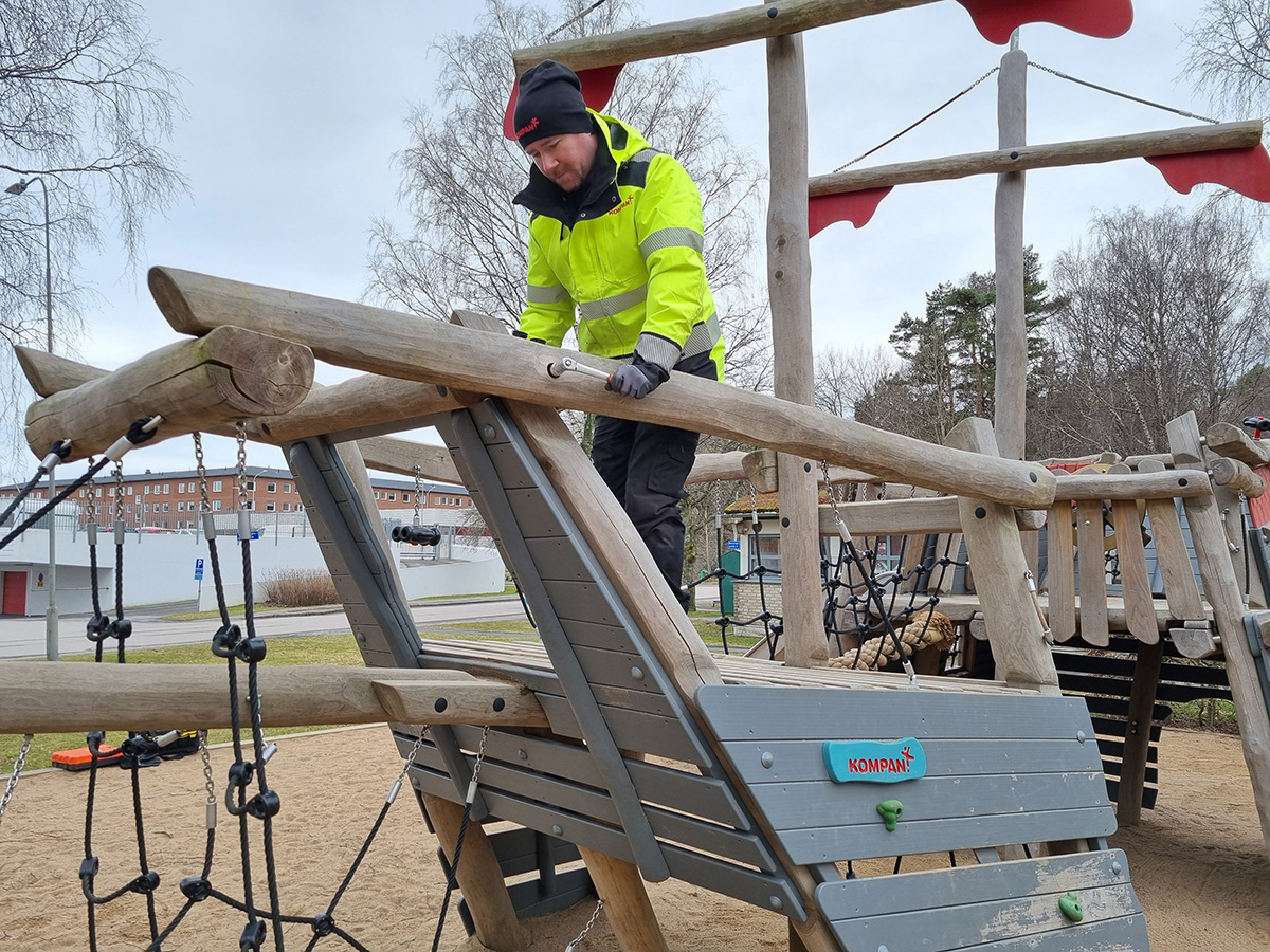 Person in neon yellow jacket climbing a wooden playground structure with ropes and sand on a cloudy day.