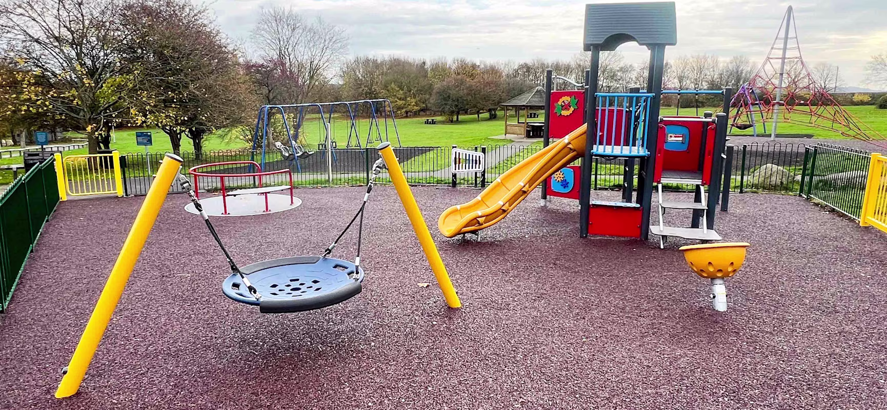 Colourful fenced playground with slide, swings and roundabout on rubber surface, set in a grassy park under cloudy sky.