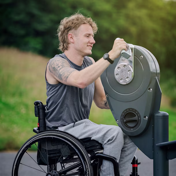 Man in a wheelchair using an outdoor upper-body exercise machine on a path with green trees in the background.