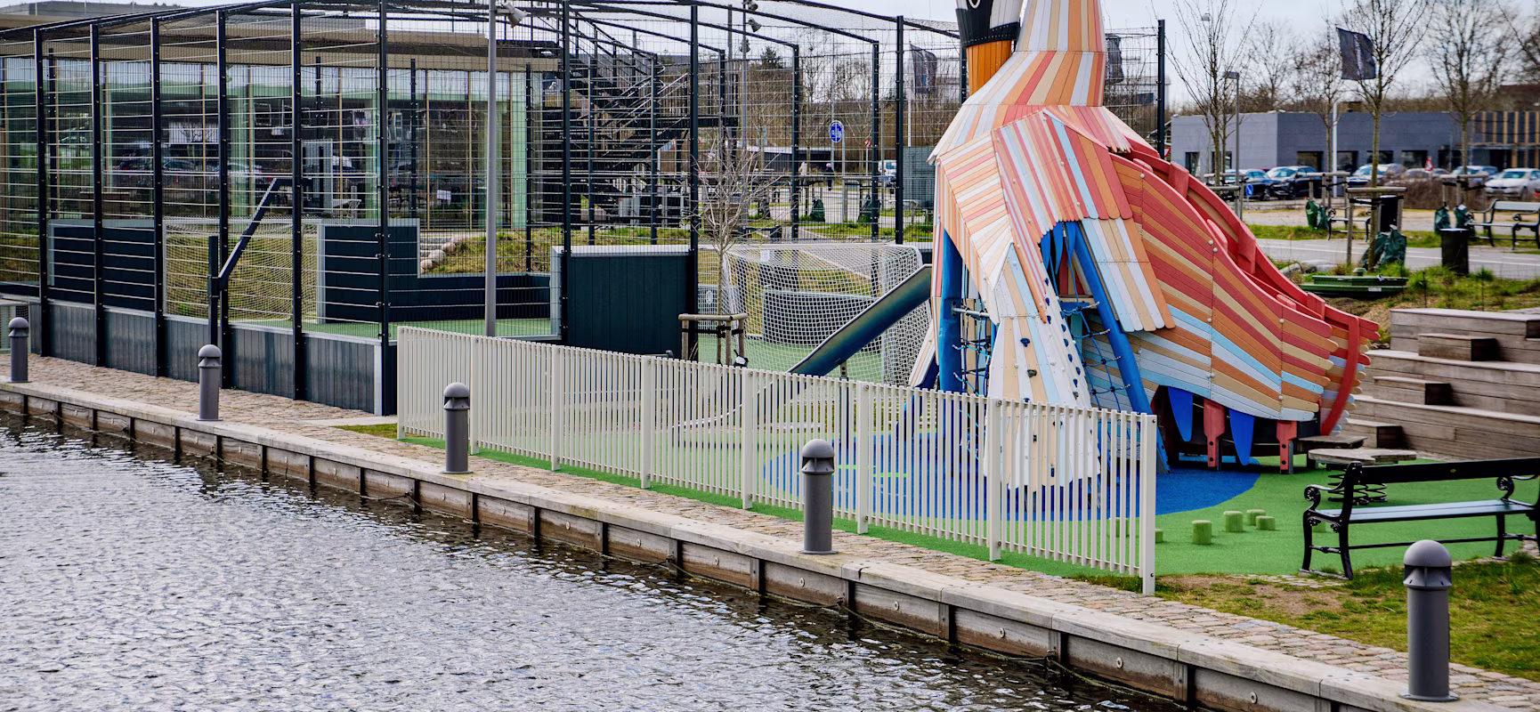 Colourful bird-shaped playground slide beside a fenced sports court near a waterside path with benches and bollards