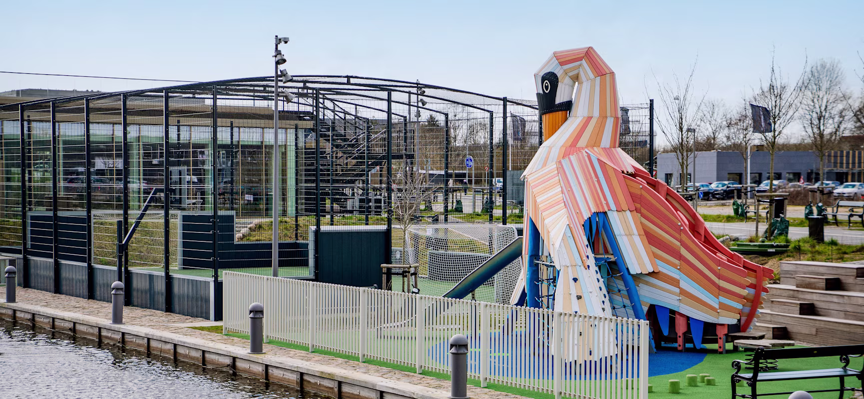 Colourful bird-shaped playground slide beside a fenced sports court near a waterside path with benches and bollards