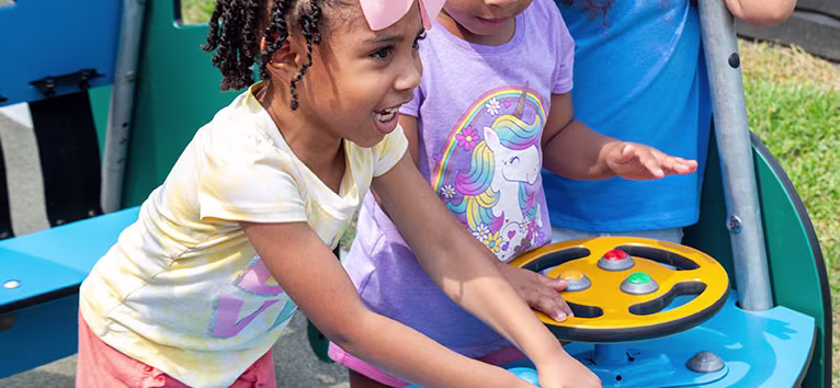 Three preschool-age children playing in a car-themed piece of early years outdoor play equipment.