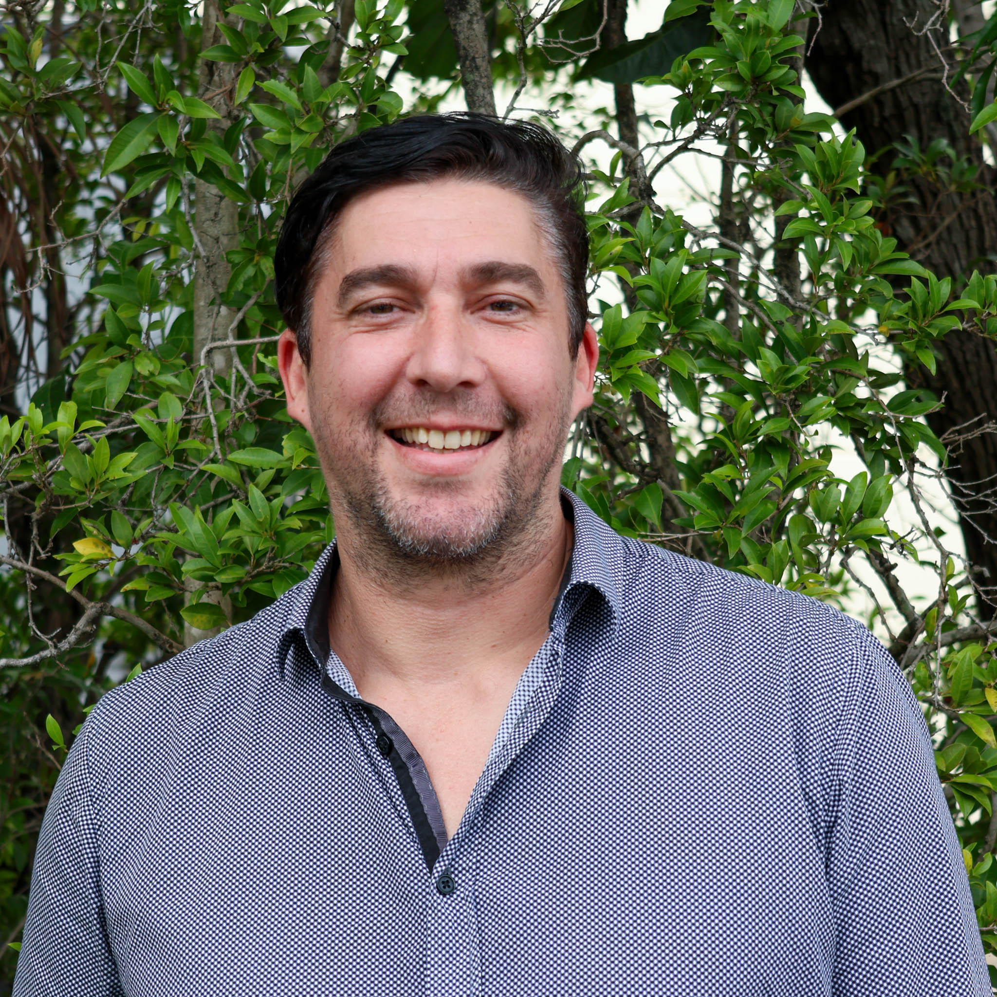Person with blurred face wearing a patterned button-up shirt, standing outdoors in front of green leafy bushes
