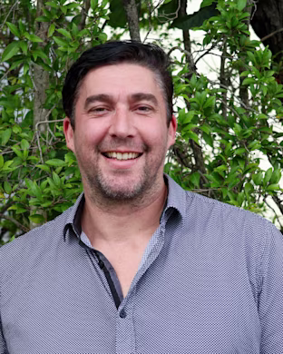 Person with blurred face wearing a patterned button-up shirt, standing outdoors in front of green leafy bushes