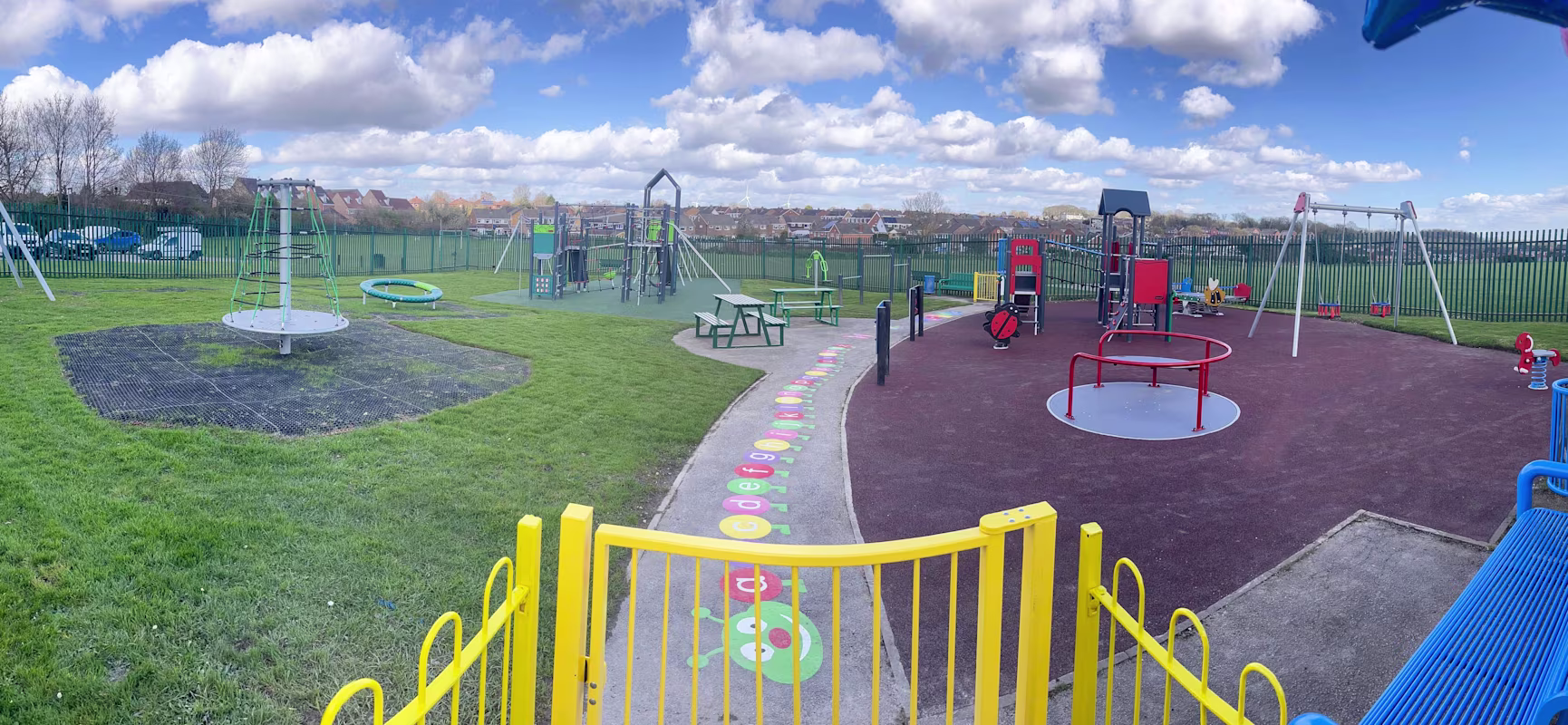 Colourful fenced playground with swings, climbing frames, roundabouts and benches on grass and rubber, under a blue sky with clouds.