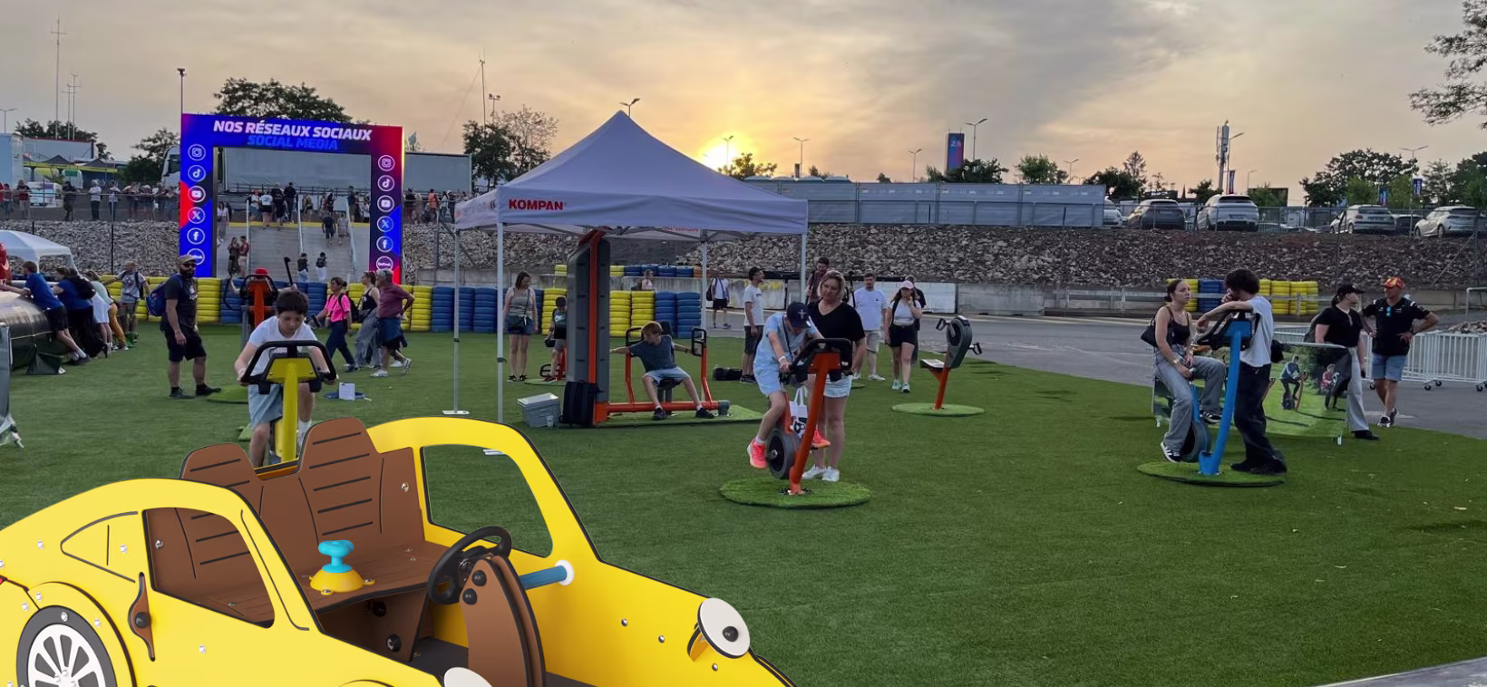 Outdoor event at sunset with people using fitness equipment on artificial grass, colorful archway, tents, and a cartoon yellow car in front