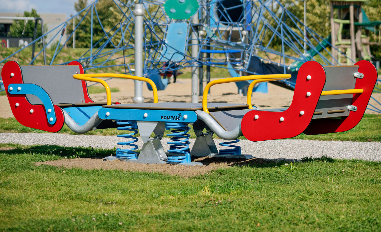 Colorful spring rocker and climbing net structure in a sunny outdoor playground with grass, sand, and trees in the background.