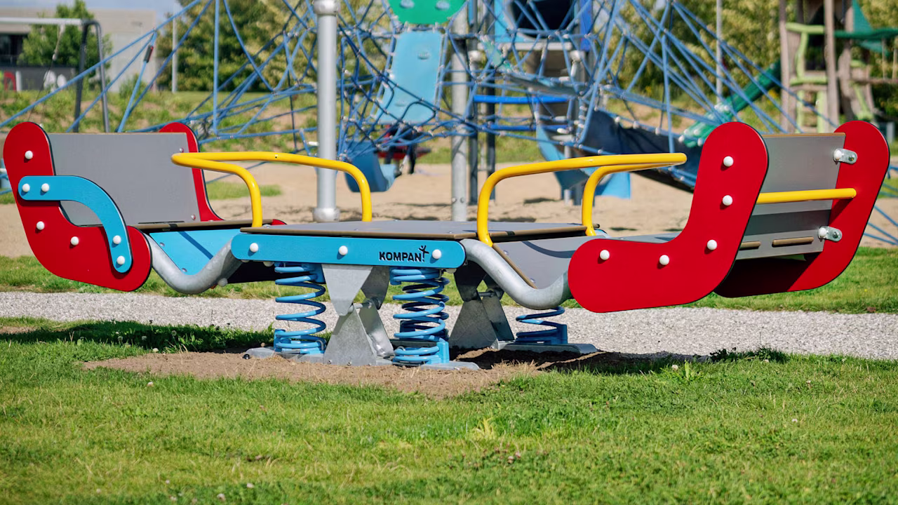 Colorful spring rocker and climbing net structure in a sunny outdoor playground with grass, sand, and trees in the background.