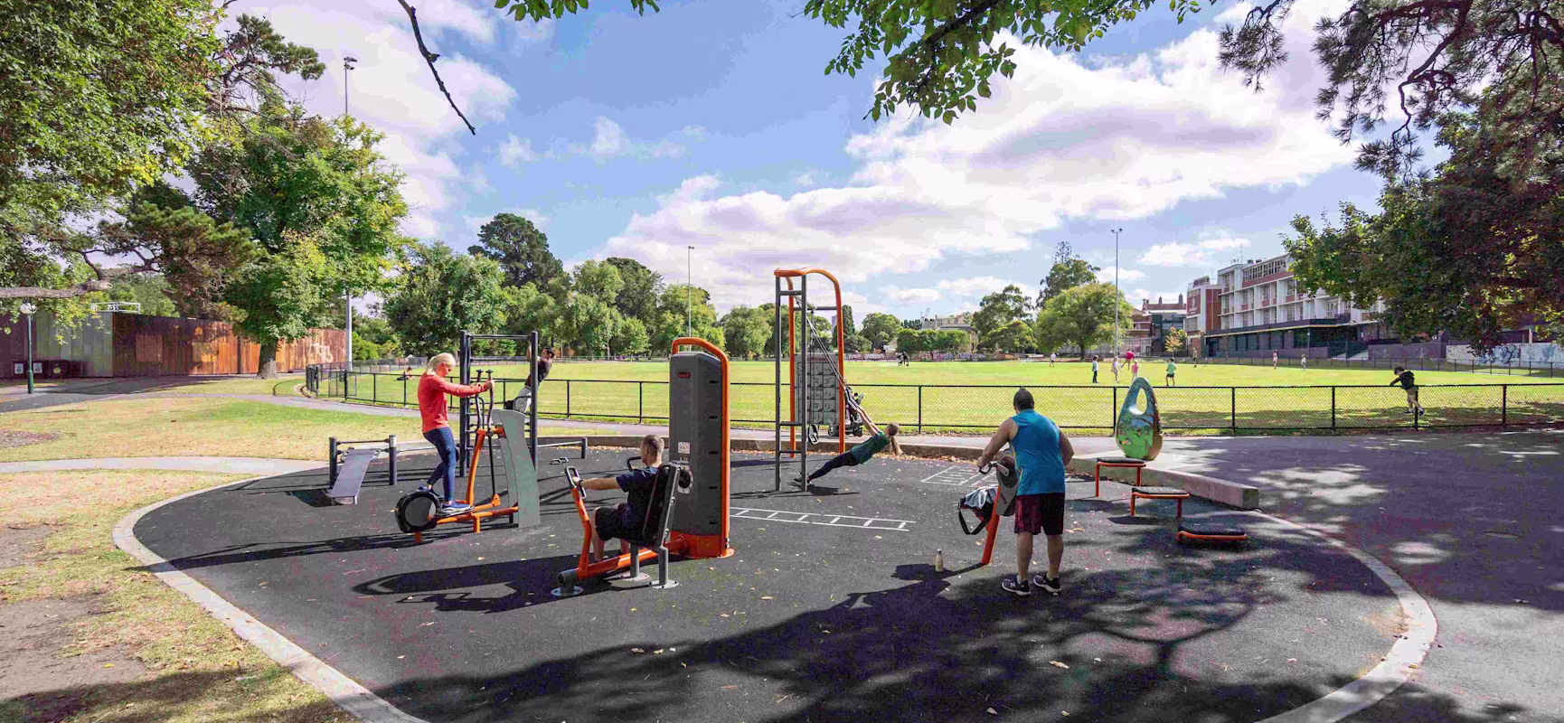 People using outdoor gym equipment beside a green sports field on a sunny day, with trees and buildings in the background