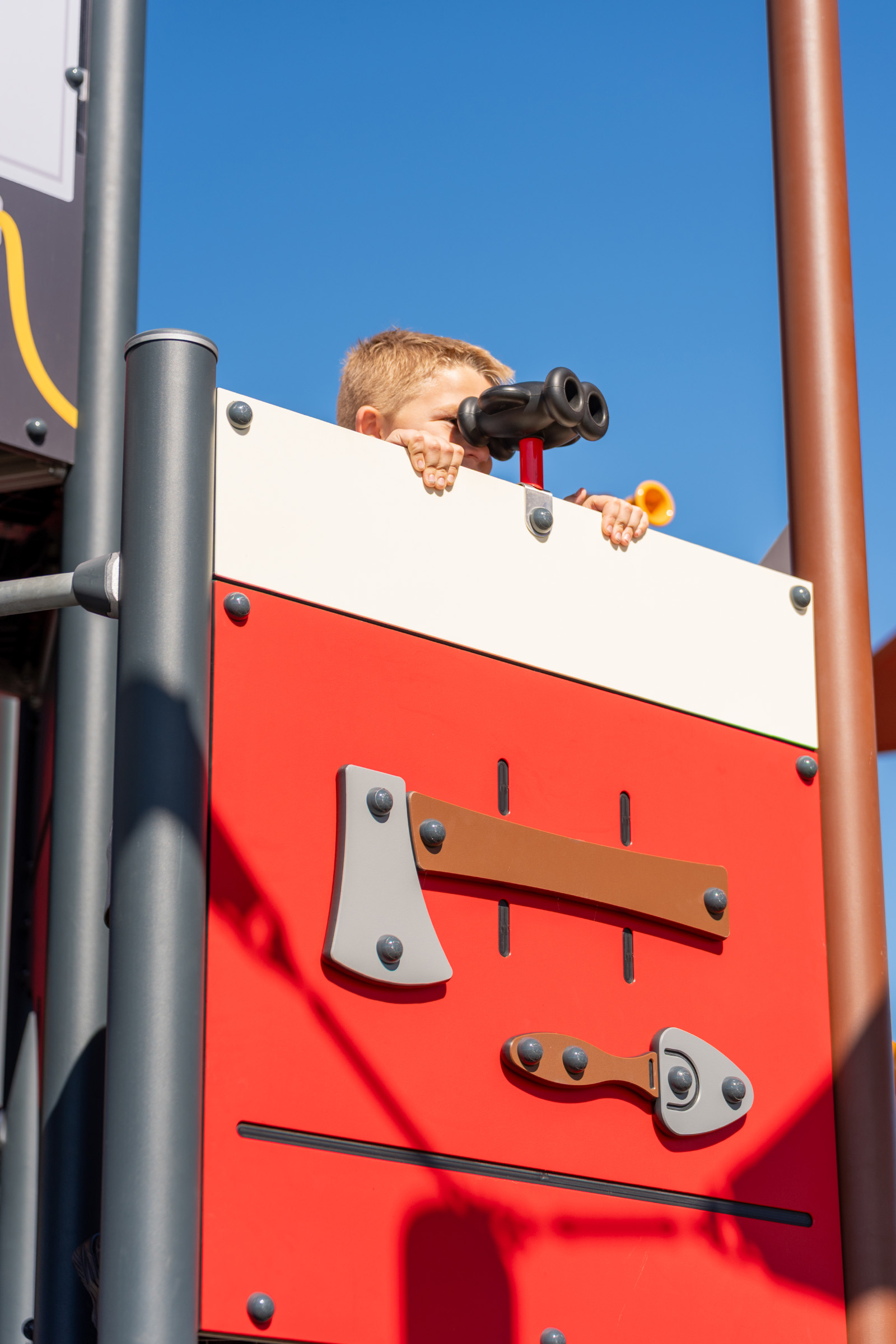 Child hidden behind red and white playground wall, peeking through toy binoculars, under clear blue sky.